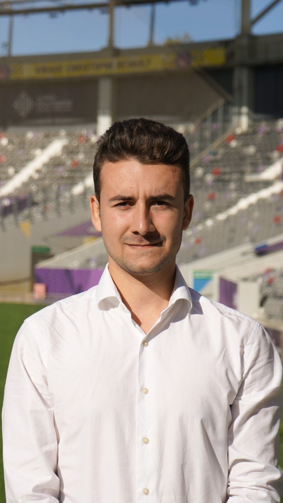 Jeune homme en chemise blanche debout dans un stade en plein air, avec des tribunes en arrière-plan.