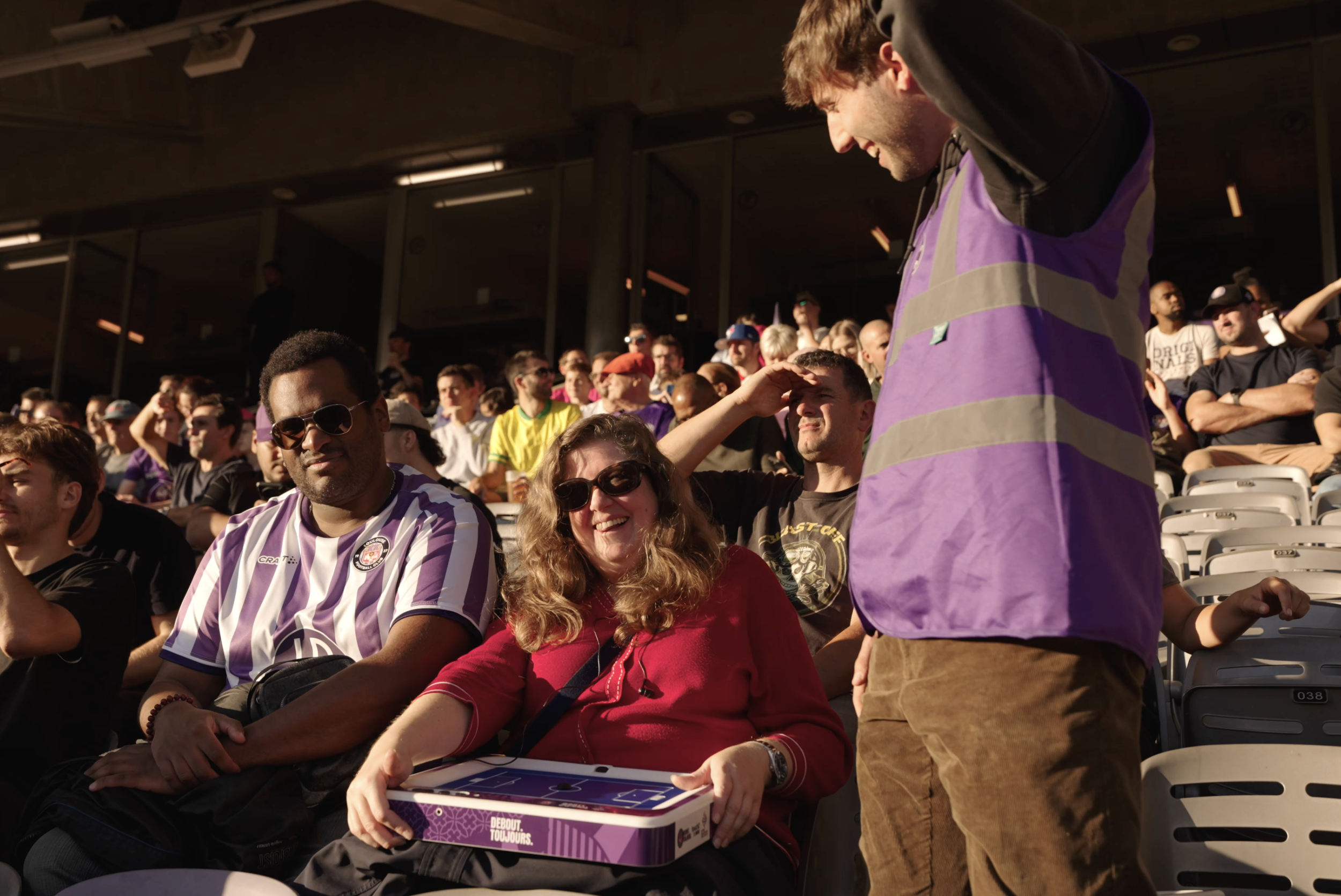 Groupe de spectateurs dans un stade, certains portant des vêtements violets et mauves, en train d'assister à un événement sportif ou de divertissement.